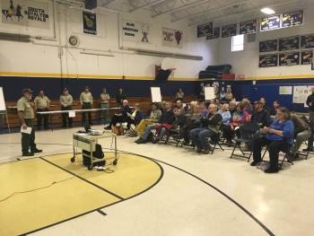 A group of people seated in a gymnasium, being spoken to by National Forest staff.