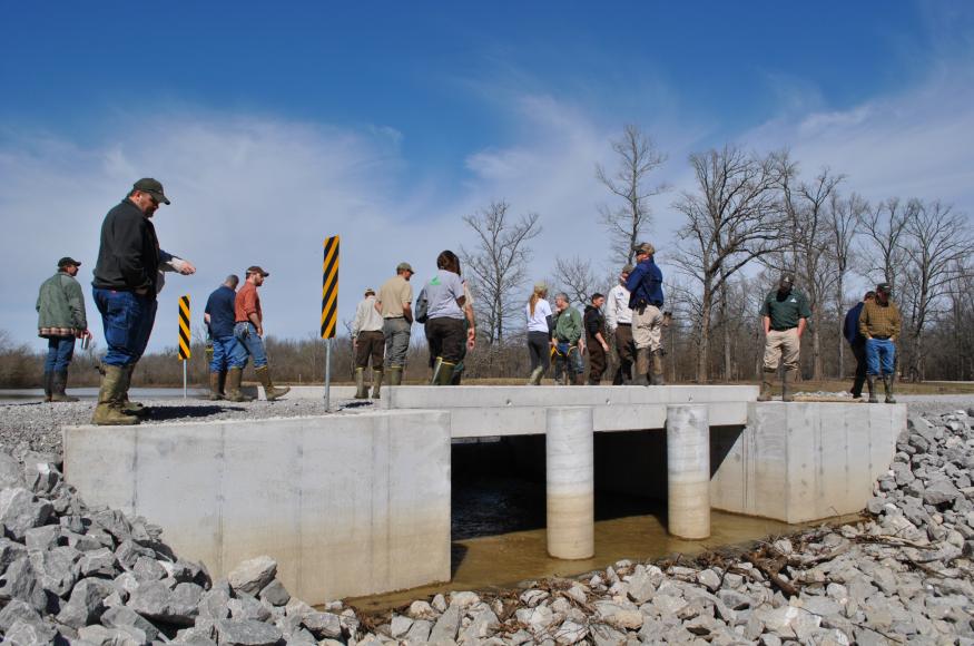 A group of people stand on a road with a box culvert underneat